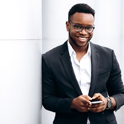 person in a suit and glasses on their phone and standing against a wall