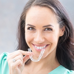 A young woman holding a clear aligner and smiling after learning about the cost of Invisalign