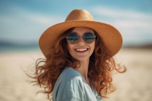 smiling woman with long curly hair wearing sunglasses and a straw hat on the beach